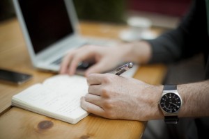 A man writes in a journal near a laptop